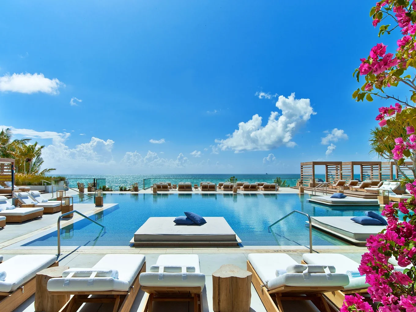 Sun-soaked outdoor pool area designed for relaxation and well-being, featuring natural wooden decking and ocean views under a clear blue sky; pink flowers at the edge add a touch of harmony—an inviting environment that supports gut health through stress reduction, fresh air, and opportunities for mindful rest.