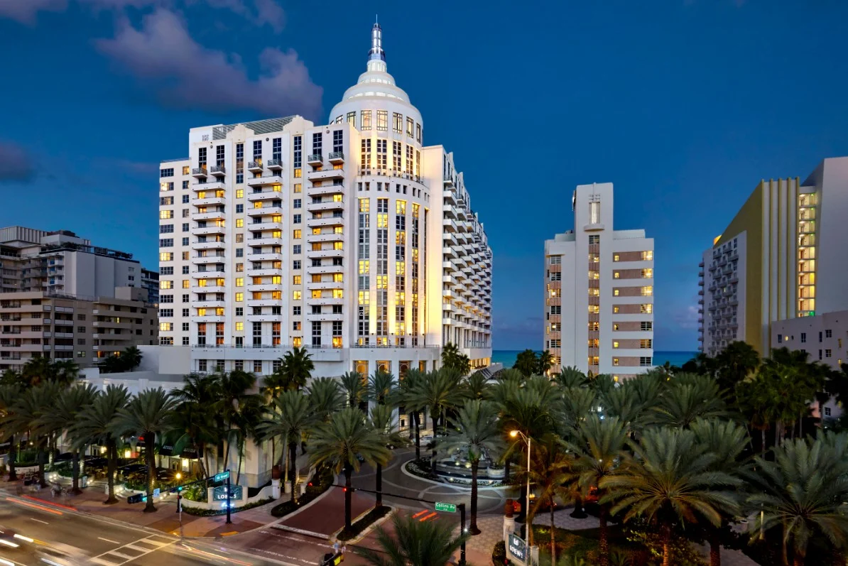 A tall, white wellness hotel—with warmly lit windows—stands among lush palm trees at dusk, offering a serene retreat for restorative gut health. Neighboring high-rise buildings hint at the vibrant city beyond, while the darkening sky sets a tranquil mood for rejuvenation and mindful nourishment.