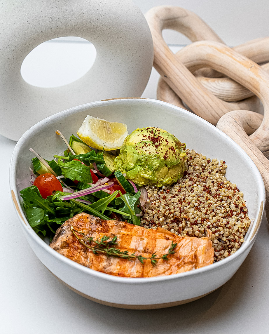 A nourishing gut-friendly bowl featuring grilled salmon, fiber-rich quinoa, creamy avocado, a variety of mixed greens for prebiotics, juicy cherry tomatoes, gut-supporting red onion, and a lemon wedge for digestion—all arranged on a white surface with modern decorative objects in the background.