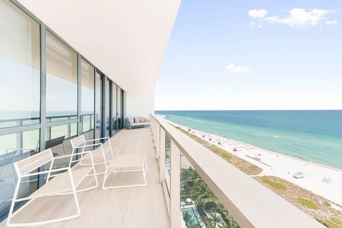A serene balcony designed for relaxation and gut health overlooks a sandy beach and the expansive ocean beneath a clear, blue sky. White chairs invite mindful moments, while large glass windows to the left bathe the space in natural sunlight, creating an environment that nurtures well-being and supports healthy digestion.